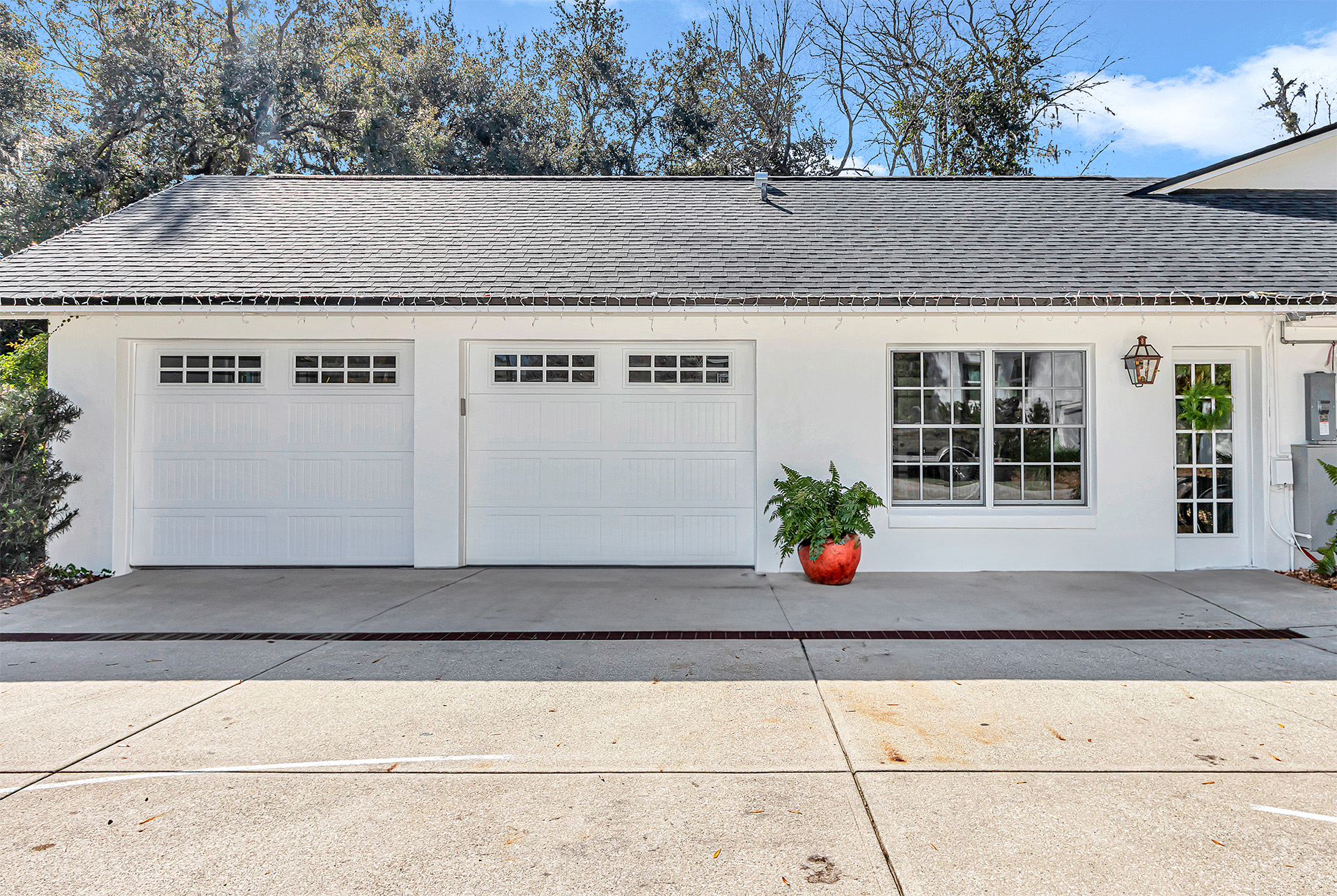 A well-maintained white garage with double doors, flanked by a decorative red planter with greenery, and adjacent to a welcoming entrance by the driveway.