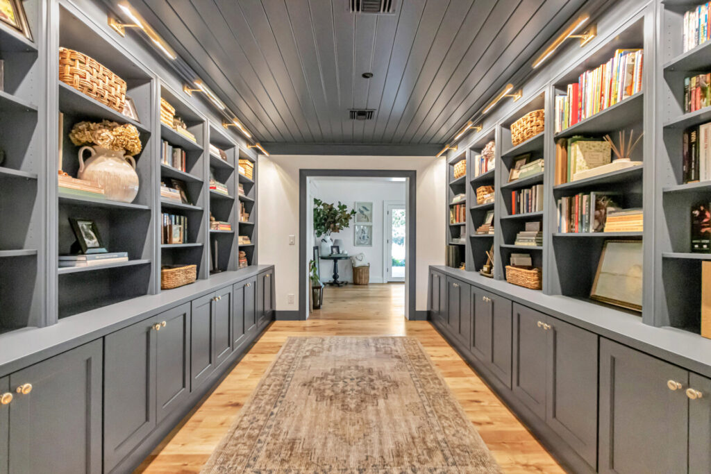 A spacious home library featuring gray built-in shelves filled with books, decorative items, and woven baskets, complemented by soft lighting and a wooden floor.