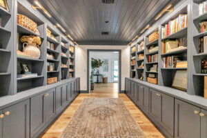 A spacious home library featuring gray built-in shelves filled with books, decorative items, and woven baskets, complemented by soft lighting and a wooden floor.