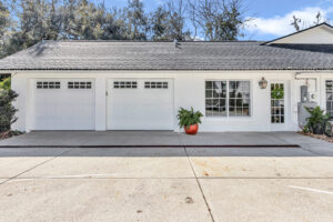 After Garage Conversion: A well-maintained white house exterior featuring two garage doors, a decorative lantern, and a vibrant planter with greenery in front, surrounded by a clean driveway and trees in the background.
