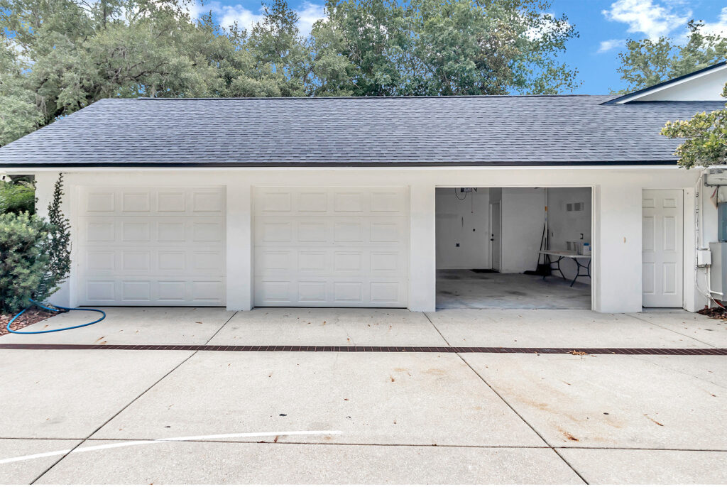 A view of a garage featuring three large white doors, one partially open, with a clean driveway and greenery in the background.