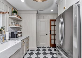 A sleek modern kitchen featuring a farmhouse sink, marble countertops, open shelving, a stainless steel refrigerator, and a checkerboard tile floor.