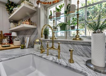 A modern kitchen featuring a white marble countertop, brass fixtures, and potted plants near large windows, creating a fresh and stylish atmosphere.